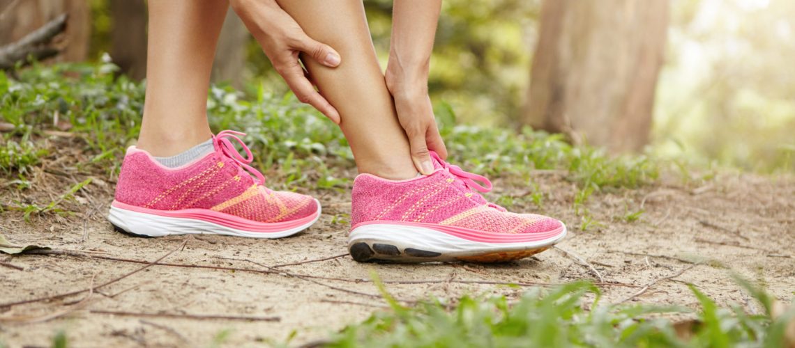 Running sport injury. Female athlete jogger wearing pink sneakers touching her twisted or sprained ankle while jogging or running outdoors. Woman runner massaging her calf muscle before workout